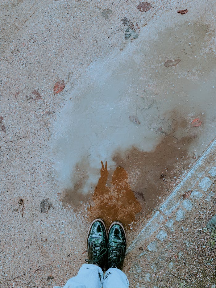 From above silhouette of anonymous person with peace sign reflected in puddle on asphalt in street in city in daytime