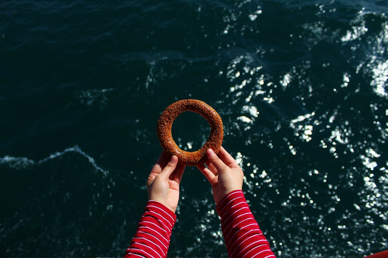 A person holds a sesame-covered bagel over the shimmering ocean on a sunny day.